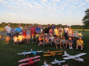 A group of people poses outdoors on a lawn surrounded by model airplanes at sunset.