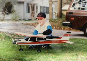 Child smiling with a red and white model airplane on grass.