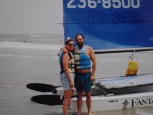Couple in life vests stands near a sailboat on a beach.