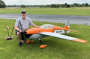 Man kneeling beside large orange and white model airplane on grass.