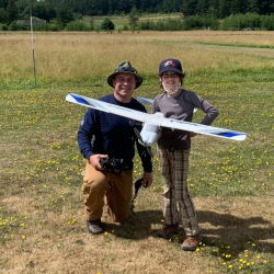 Person kneeling with a child holding a model airplane on a grassy field.