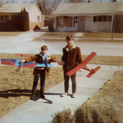 Two children holding colorful model airplanes on a suburban sidewalk in front of houses.