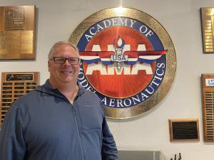 Man smiling in front of Academy of Model Aeronautics logo on wall.