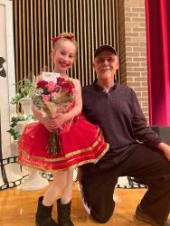 Young girl in red dance costume holding flowers, smiling with a man kneeling beside her indoors.