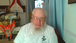 Senior man with white beard and glasses, wearing a white T-shirt, sitting indoors.