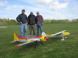 Three people stand on grass beside large remote-controlled airplanes.