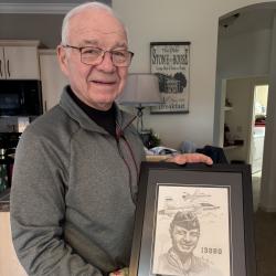 Elderly man smiling, holding a framed sketch of himself in military attire with aircraft in background.