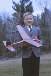 A boy smiling, holding a model airplane, stands outdoors in a suit and tie.