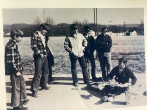 A group of people stand around someone seated, looking at a model airplane outdoors.