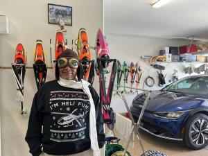 Man wearing ski gear and goggles in a garage with skis and a blue car.