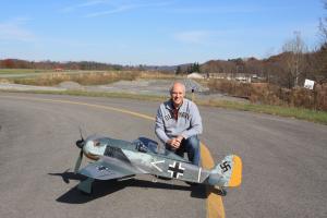 Man kneeling beside a large model aircraft on a road under a clear blue sky.