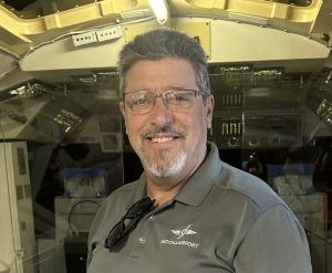 An older man smiles in an aircraft cockpit, wearing a polo shirt and glasses.