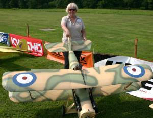 Woman with model biplane, camouflage paint, in a grassy field.