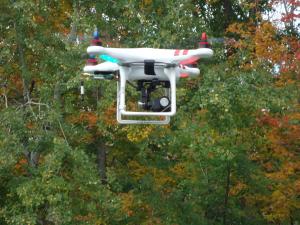 A white drone flying in front of colorful autumn trees.