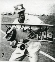 Man holding a model biplane on a grassy field, wearing a cap and shirt.