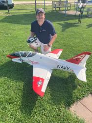 Man crouching beside large model Navy jet on green grass.