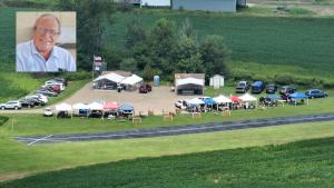 Rural event with tents and booths near parked cars. Inset of smiling man.