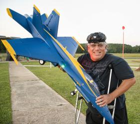 Man smiling, holding a large model blue and yellow jet, standing on grass with crutches.