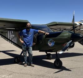 Man in blue shirt standing next to a green and black aircraft on a sunny day.