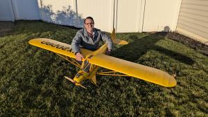 Man smiling with a large yellow model airplane on grass.