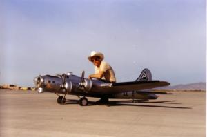 Man sitting on miniature B-17 model airplane on a runway, wearing a cowboy hat.