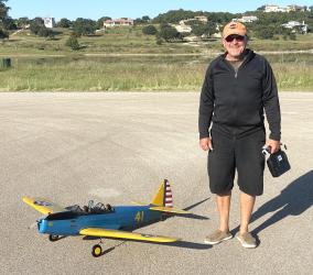 Man smiling next to blue and yellow model airplane on an airfield.