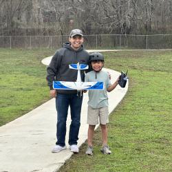 Father and son with model airplane and remote control, smiling on a path in a grassy park.