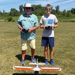 Two people standing outside holding remote controls, with a model plane on grass.
