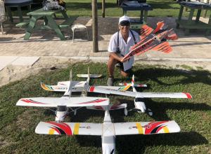 Person smiling, kneeling on grass with model airplanes in front, holding orange and black plane.