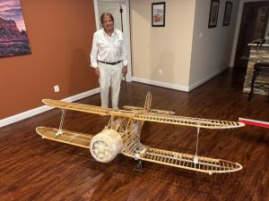 Man standing behind large wooden model airplane in a room with wooden floor.