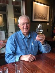 Elderly man in a blue shirt, smiling, holding a wine glass in a cozy restaurant.