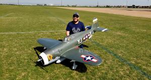 Man kneeling on grass beside a large model fighter plane.