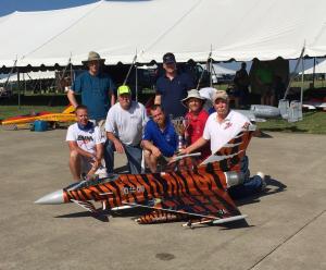 Men posing with a tiger-striped model jet, holding a trophy, in front of a tent.