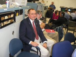 A man in a suit sits in a control room, surrounded by people at computers.