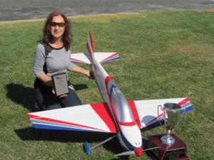 Woman with remote control plane, holding a plaque beside a trophy on grass.