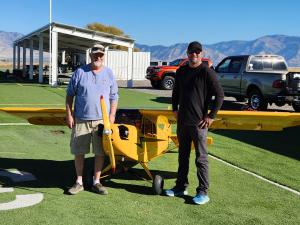 Two men stand beside a yellow model airplane on a grassy field, with mountains in the background.