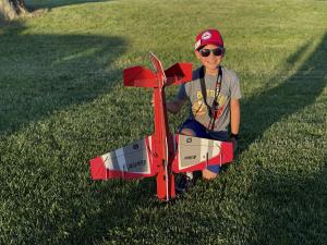 A child in a red hat smiles, kneeling beside a red model airplane on grass.