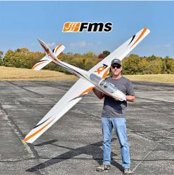 Man holding large model plane on an open airstrip, FMS logo above.