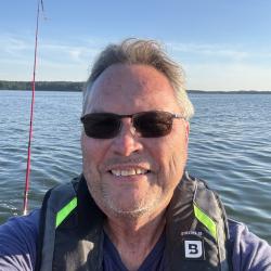 Man with sunglasses smiling on a boat with a fishing rod, calm water in the background.