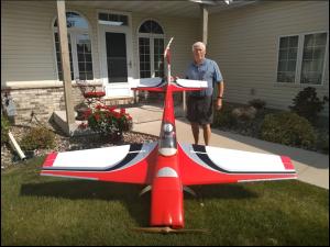 Large red model airplane on grass in front of a house, man standing beside it.