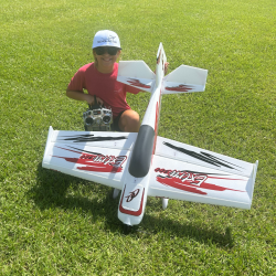 Child kneeling on grass, smiling beside a large white and red model airplane.