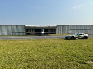 Airplane parked in front of a hangar, with a sports car nearby on a grassy lawn.