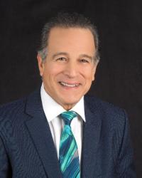 Smiling man in a blue suit with a teal tie against a black background.