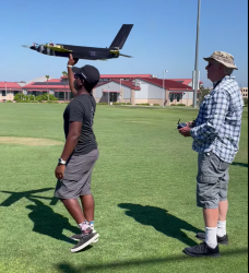 Man in black launches model plane; another observes, holds remote, both on sunny field.