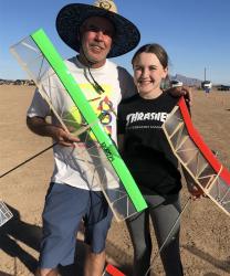 Man and woman smiling, holding model planes, standing on sunny desert ground.