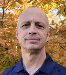Bald man in a navy shirt, standing against autumn leaves.