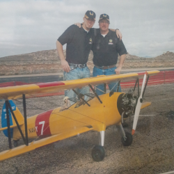 Two men in matching hats and shirts standing on an airstrip with a yellow model biplane.