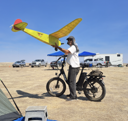 Man holding large model airplane next to an electric bike in a field under a blue sky.