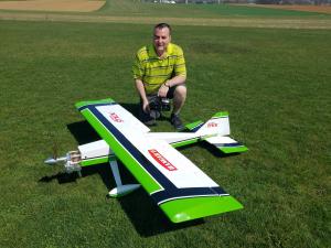 Man with remote control kneeling beside a large green and white model airplane on grass field.