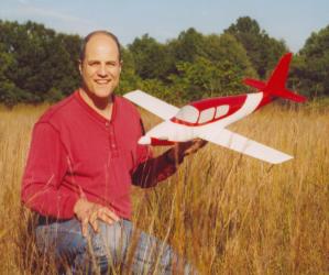 Man in red shirt holding a model airplane, sitting in a field of tall grass.